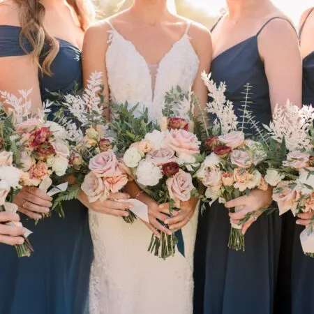 Bride and bridesmaids holding bouquets. Bridesmaids are wearing navy blue dresses. Bouquet has roses and wildflowers.